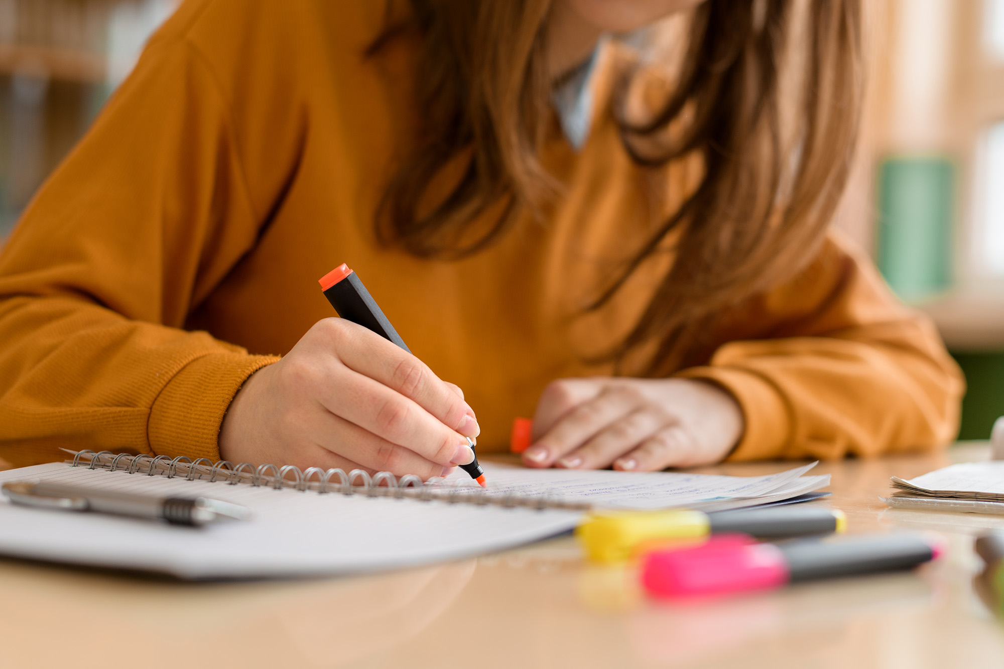 A person wearing an orange sweater writes in a notebook, honing their literacy skills. Highlighting pens are on the table.